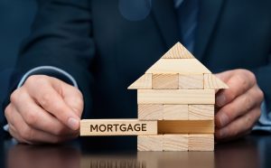 Mortgage concept. Financial agent complete wooden model of the house with last piece with text mortgage. Wide banner composition with bokeh background.